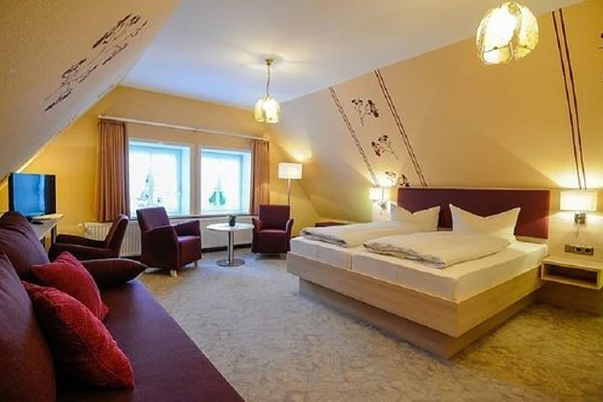 Une chambre d’hôtel avec un grand lit, un canapé, un bureau et une chaise. Il y a des fenêtres avec des rideaux, une lampe et un luminaire au plafond.