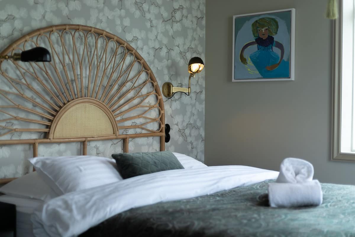 A bed with a rattan headboard, two wall-mounted lamps, a painting, and a window with daylight.