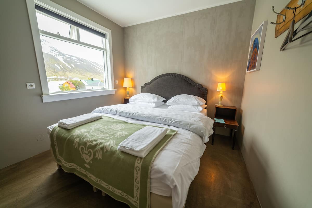 A neatly made bed with a green blanket, two bedside lamps, and a window with a view of a mountain.