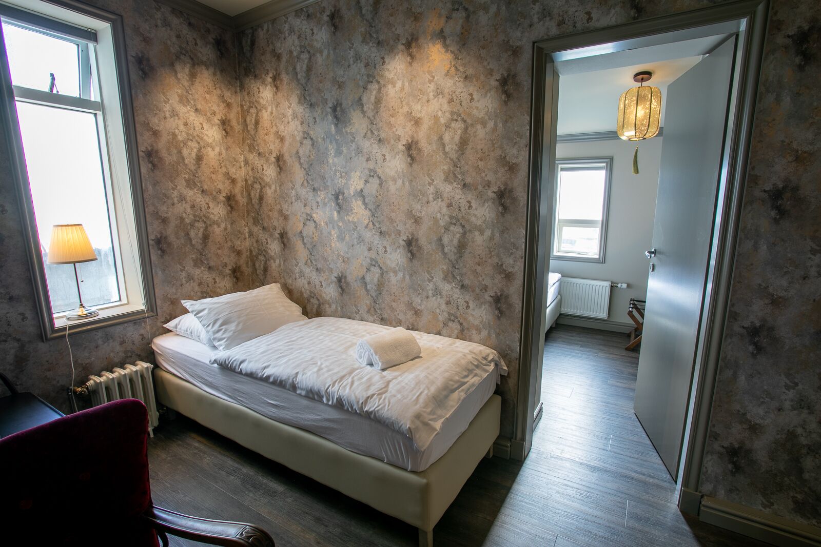 A bedroom with a bed, a chair, a radiator, and a view of another room through an open door.