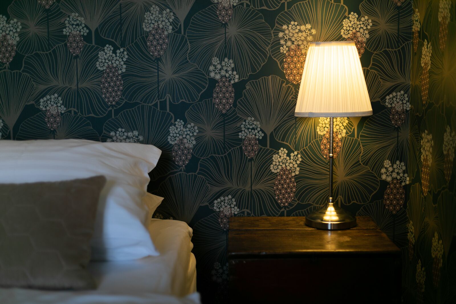 Cama bem arrumada com travesseiro, mesa de cabeceira com abajur e parede com padrão floral no papel de parede.