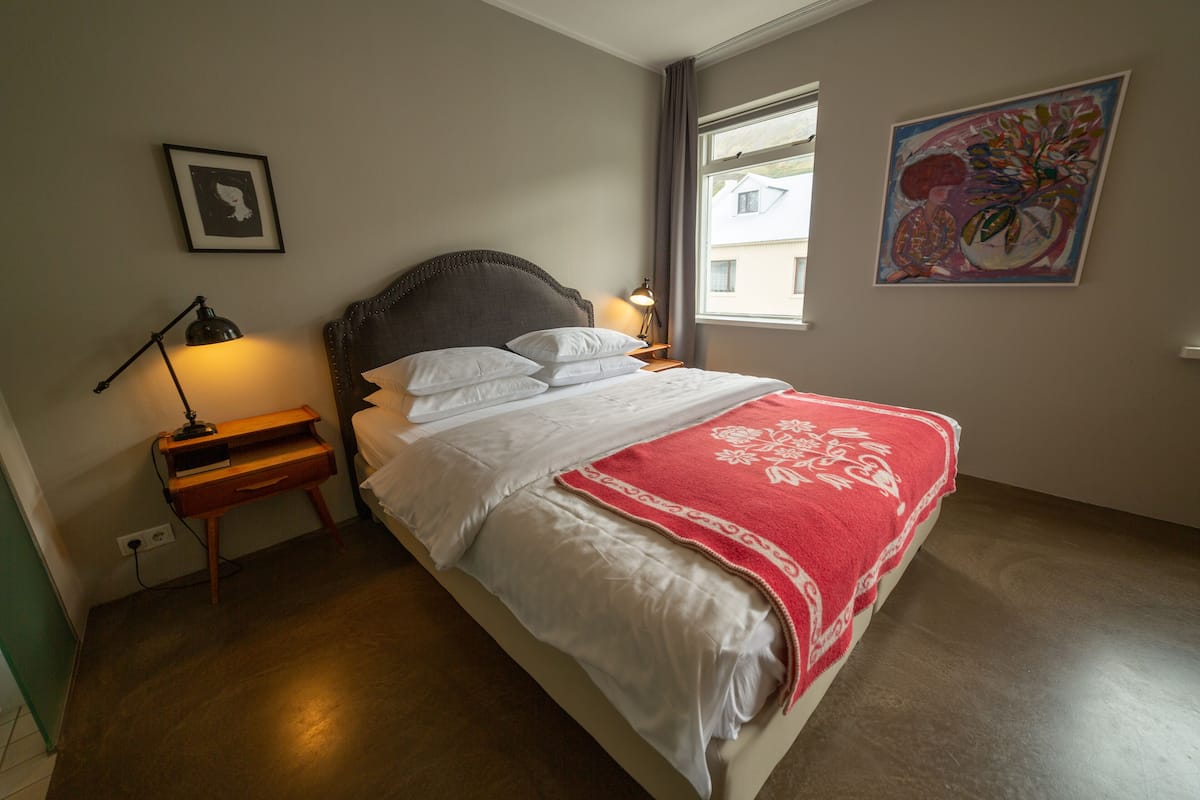 A neatly made bed with white linens and a red blanket, a bedside table with a lamp, and a painting on the wall.