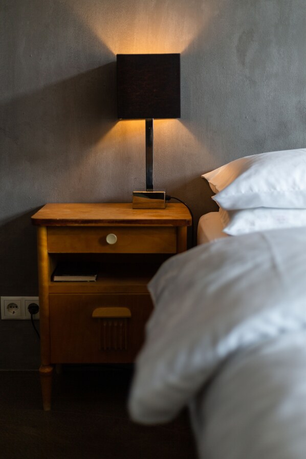 Una mesita de noche de madera con una lámpara, una cama con ropa blanca y un enchufe de pared.