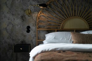 Ein Bett mit weißen Bettwäsche und einer braunen, gemusterten Decke, ein Weiden-Kopfstück, eine Wandlampe und ein kleiner, schwarzer Nachttisch mit einer Schublade.