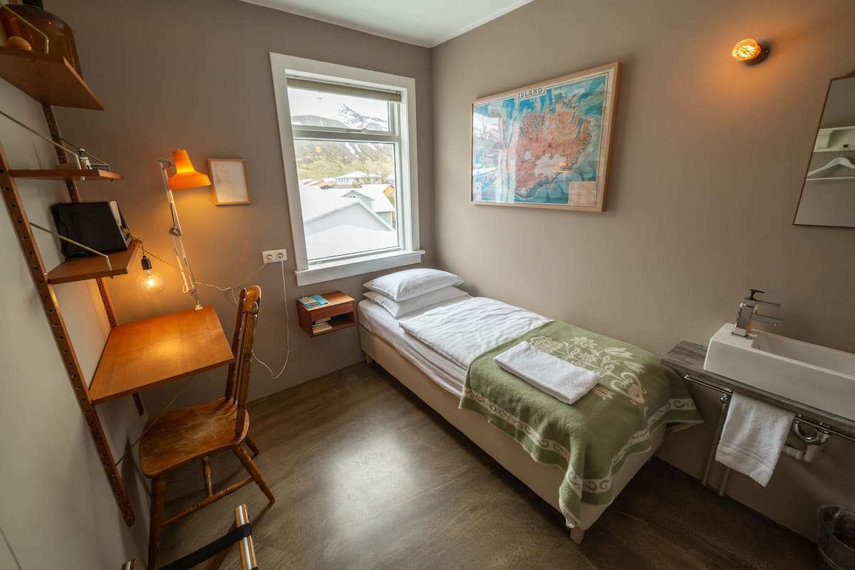 A small, well-lit room with a bed, a desk, a chair, a map on the wall, and a window with a view of a building outside.