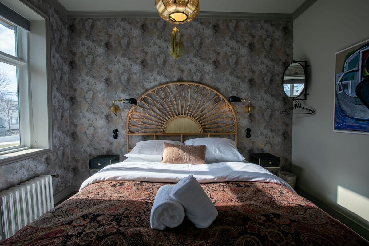 A bedroom with a large bed, a headboard with a sunburst design, two bedside lamps, a radiator, and a window with a view of the outside.