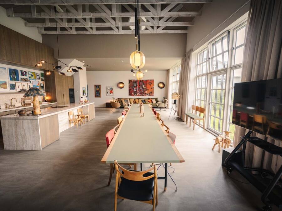 Een ruime woonkamer met een lange eettafel, diverse zithoekopstellingen en een groot raam dat voor natuurlijk licht zorgt.