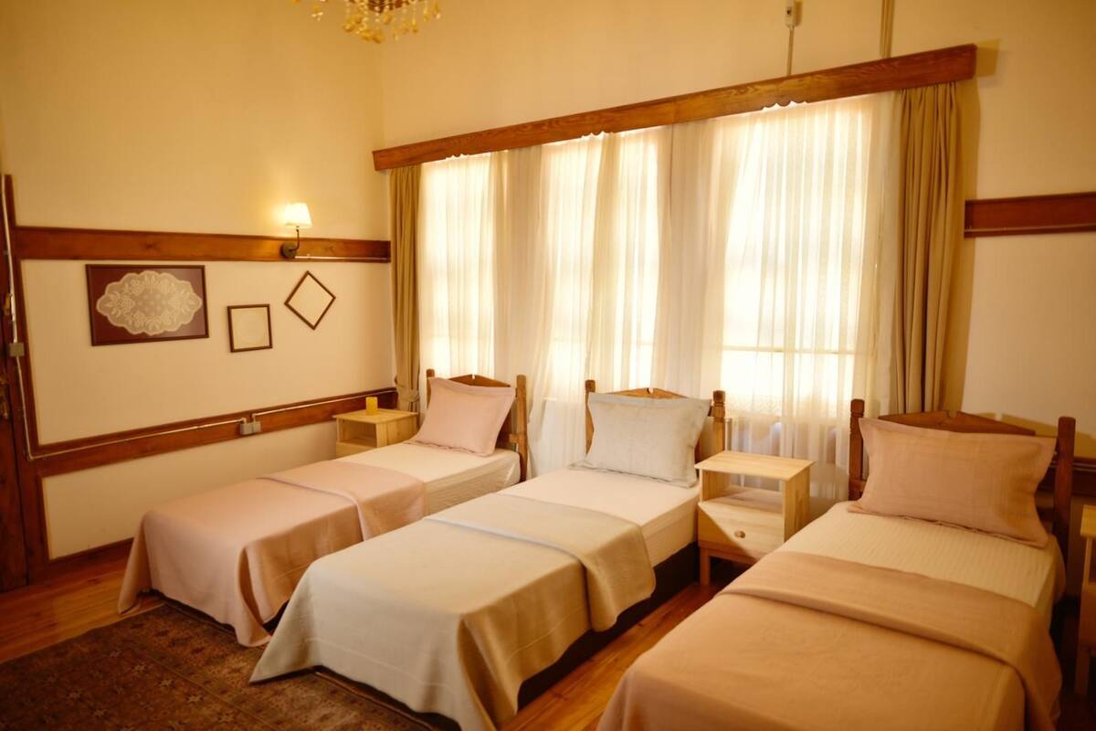A hotel room with two single beds, a wooden headboard, a nightstand with a lamp, and a large window with curtains.