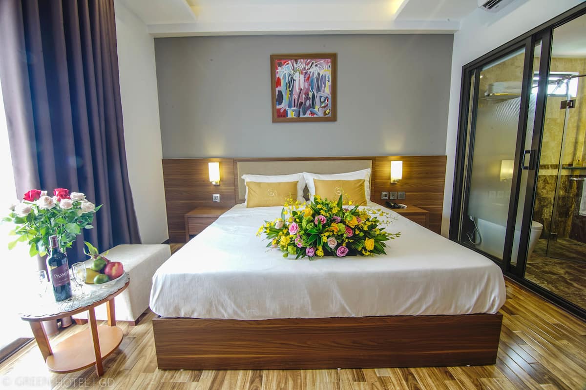 A hotel room with a large bed, a bedside table with a vase of flowers, a bottle of wine, and a glass.