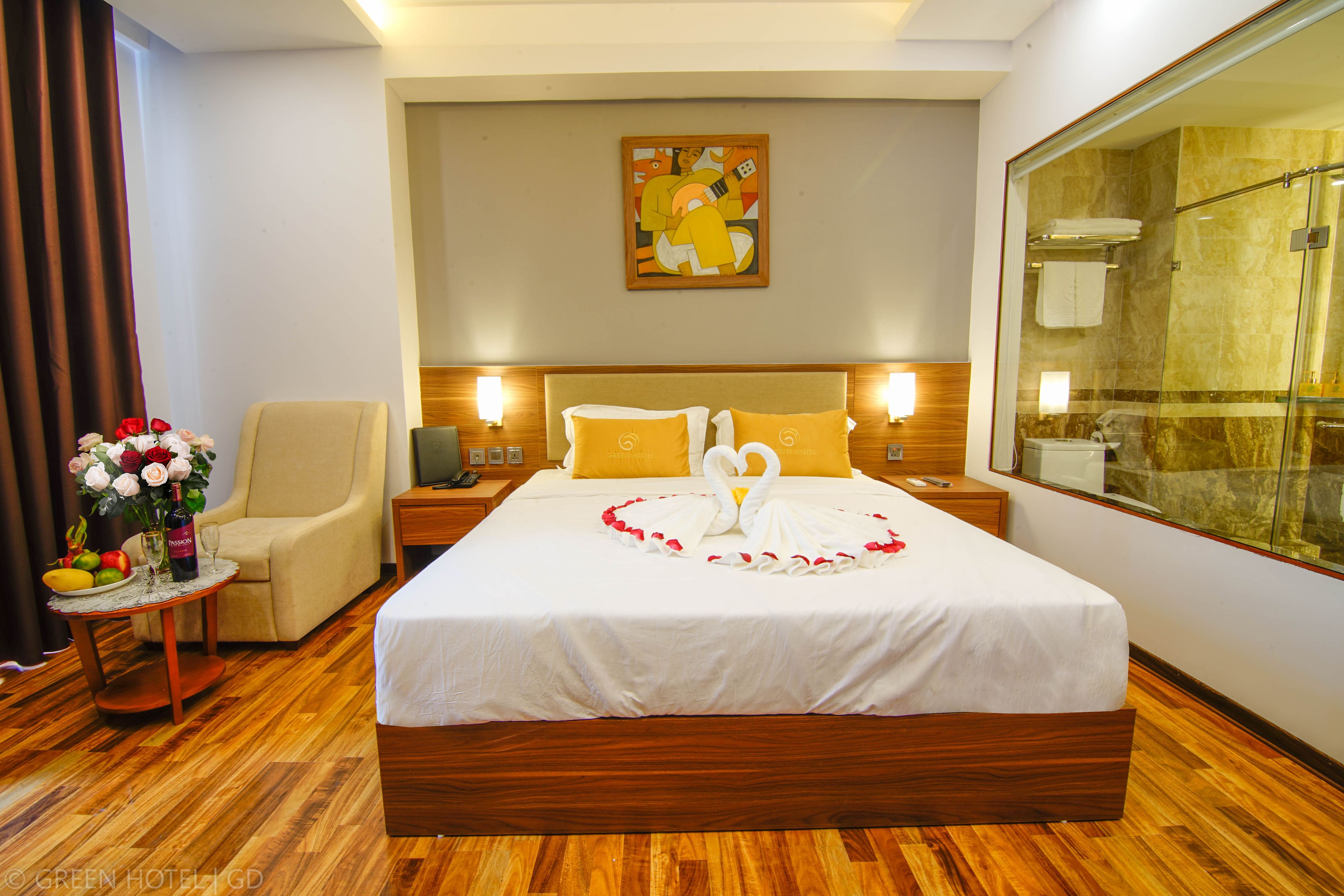 Une chambre d’hôtel avec un grand lit, un coin salon avec un fauteuil, une petite table agrémentée de fleurs et un tableau au mur.