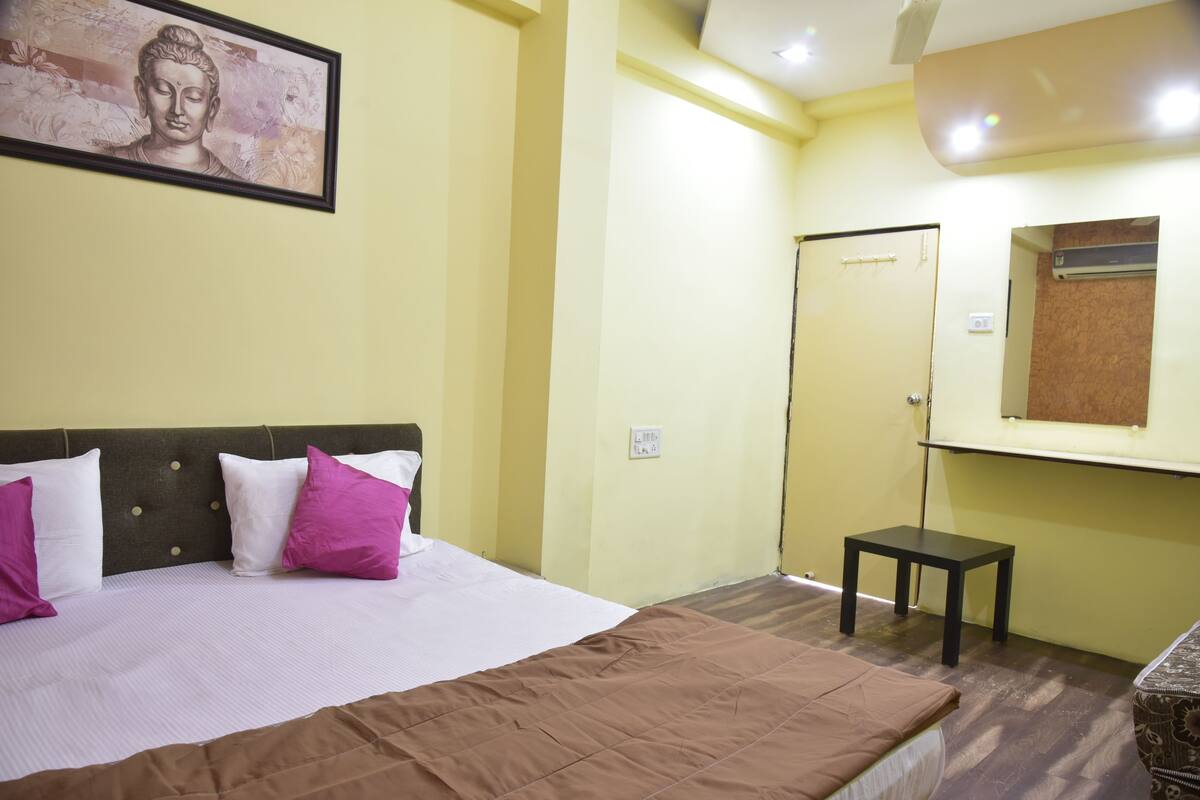 Una habitación de hotel con cama, almohadas, una mesita, una puerta y un aire acondicionado de pared.
