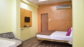 A small, compact room with a single bed, a wooden door, an air conditioning unit, a mounted television, and a small table. - Hotel Drishti Inn (Nagpur)