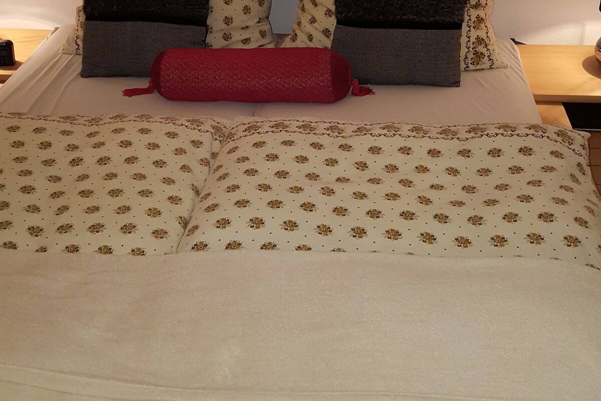 A neatly made bed with floral patterned bedding, two pillows, and a red accent pillow. A framed landscape painting hangs above the bed.
