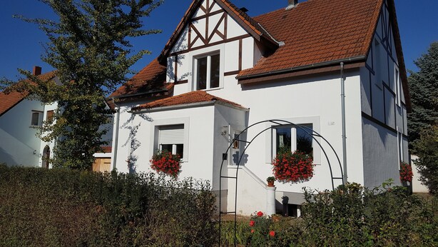 A white house with a red-tiled roof, a visible chimney, and flower boxes with red flowers. - BellaCaro (Loehne)