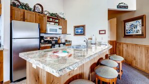 A kitchen with wooden cabinets, a stainless steel refrigerator, a microwave, a sink, and a granite countertop. There are three bar stools and a framed picture on the wall. - Jackpine 8021 (Keystone)