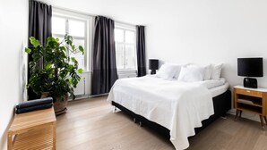 A modern bedroom with a large bed, a bedside table with a lamp, a potted plant, and a window with curtains. - Dinesen Collection Condo by Royal Theatre (Copenhagen)