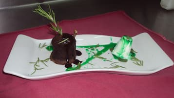 A square white plate with a dark brown dessert, green sauce, and a green cube on a red cloth.