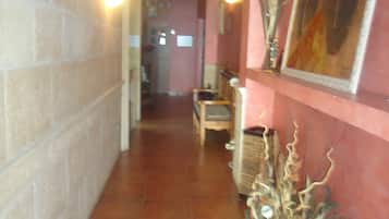 Un pasillo con suelo de baldosas, un candelabro, apliques de pared, un cuadro enmarco y una planta decorativa.