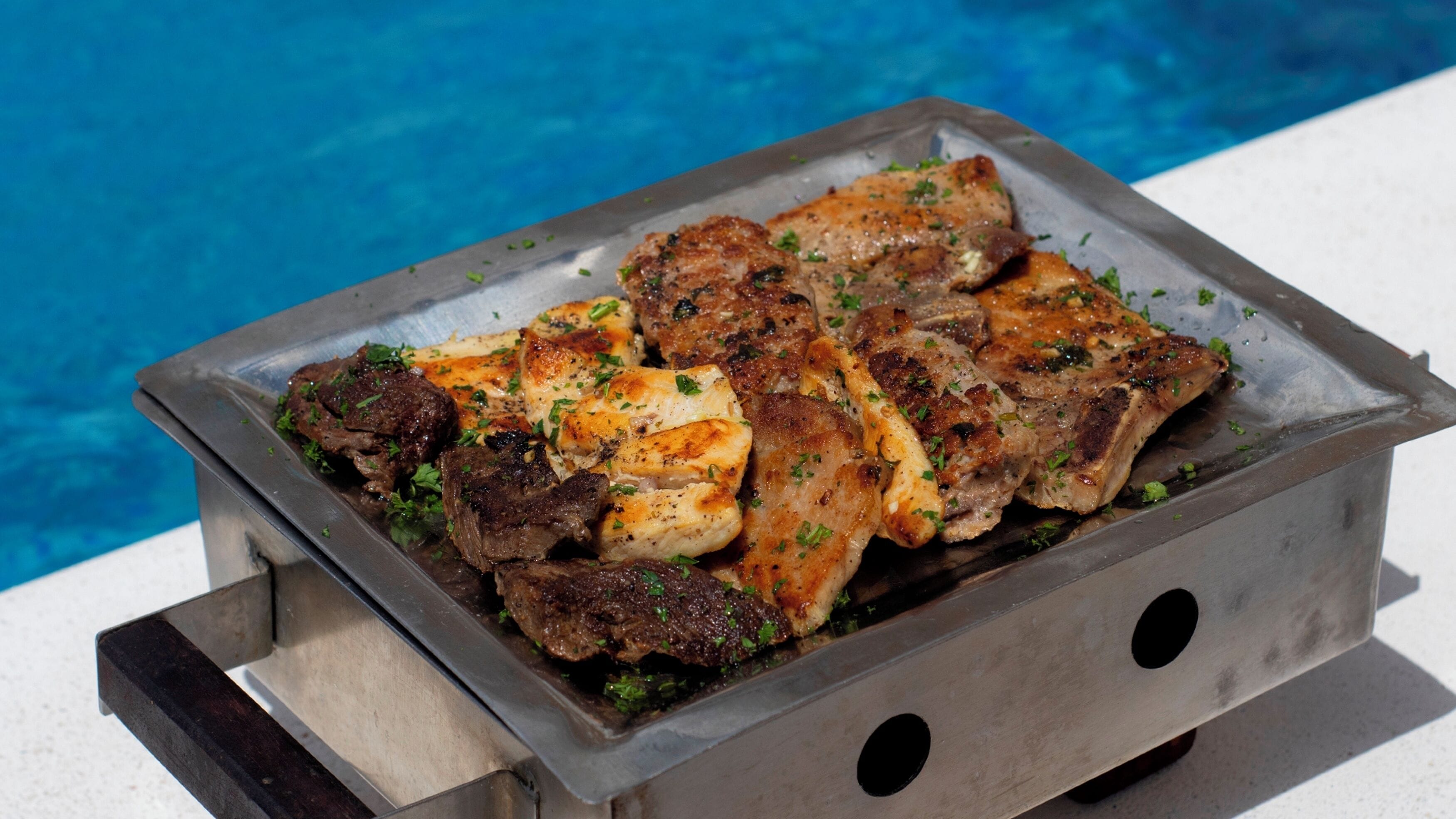 Ein Metallgrill mit gegrilltem Fleisch und Gemüse neben einem Pool.