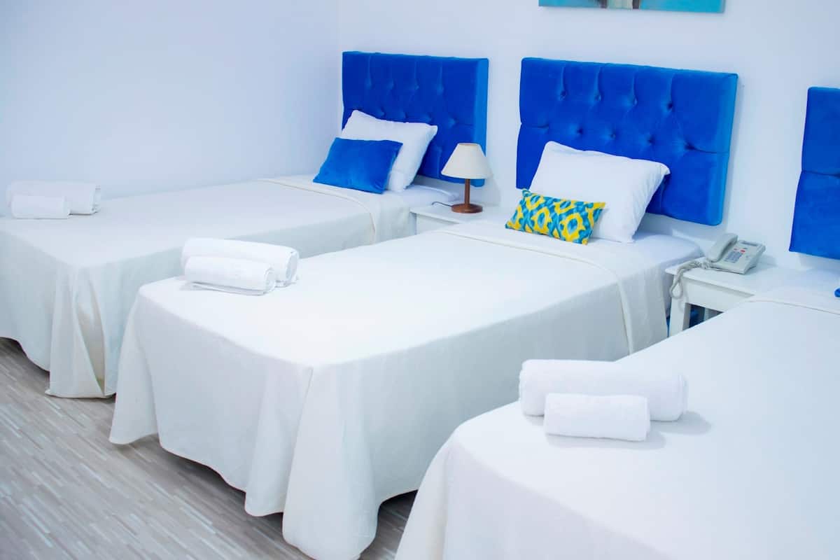 A hotel room with three single beds, each with a blue headboard and white bedding. There is a bedside table with a lamp and a phone on one of the beds.
