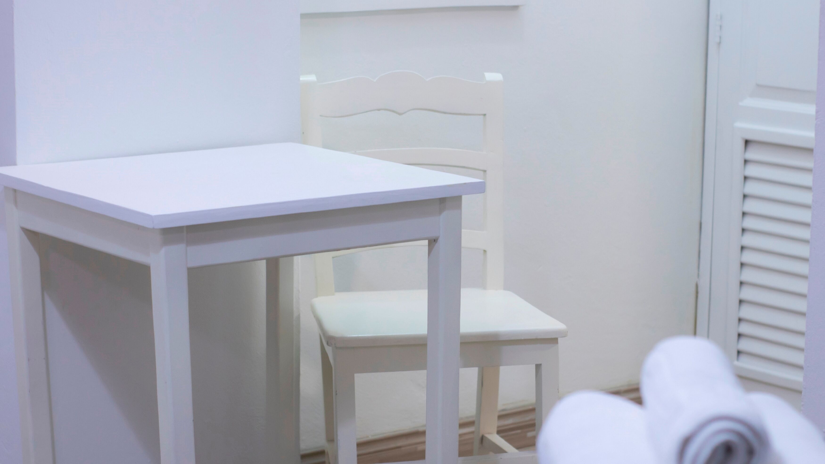 Een witte tafel en stoel in een kamer met witte wanden en een raam met zonwering.
