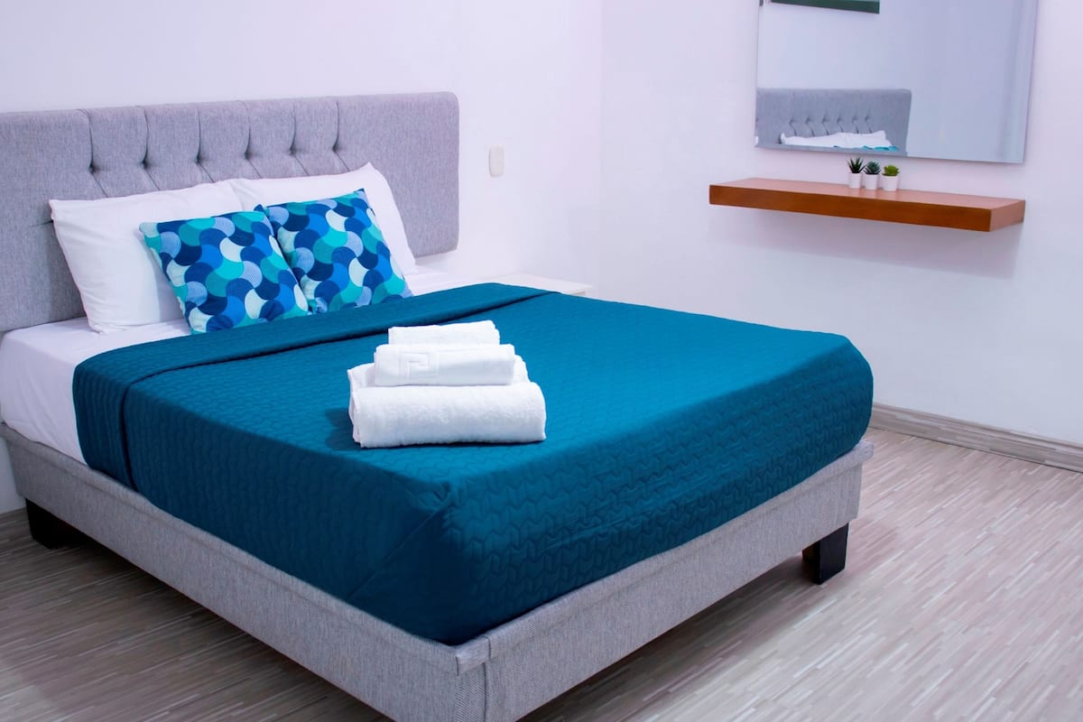 A neatly made bed with a grey upholstered headboard, white and blue patterned pillows, and a teal bedspread. There is a wooden shelf with small potted plants on the wall above the bed.