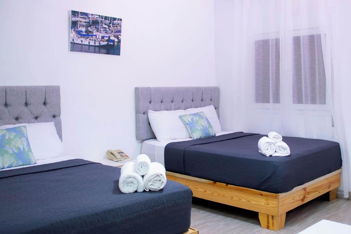 A hotel room with two beds, each with a grey headboard and a dark blue bedspread. There is a wooden bench at the foot of one bed, and a telephone on the nightstand.