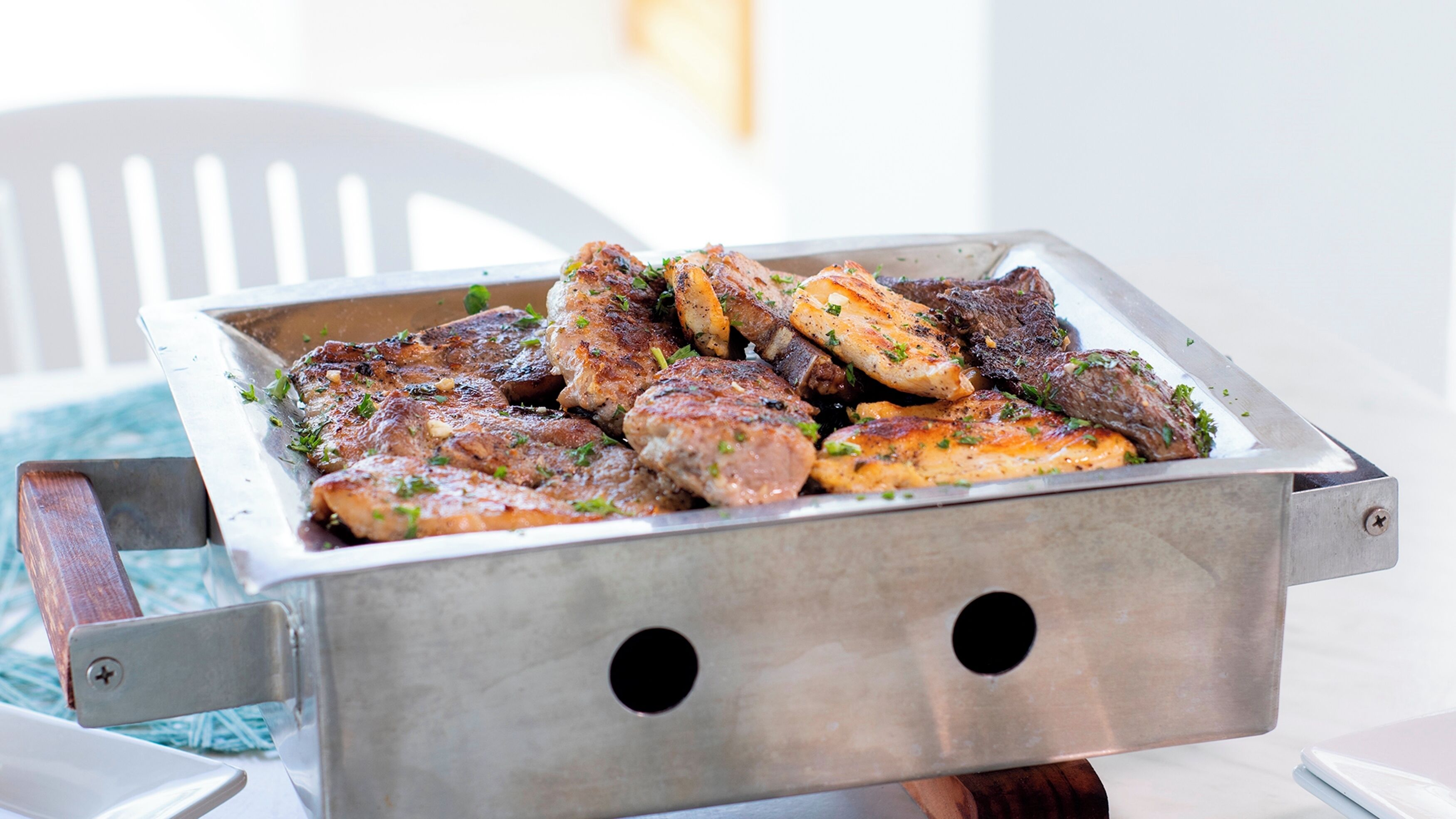 Un plateau en métal contenant de la viande et des légumes grillés, posé sur une table. On aperçoit une chaise blanche à l’arrière-plan.