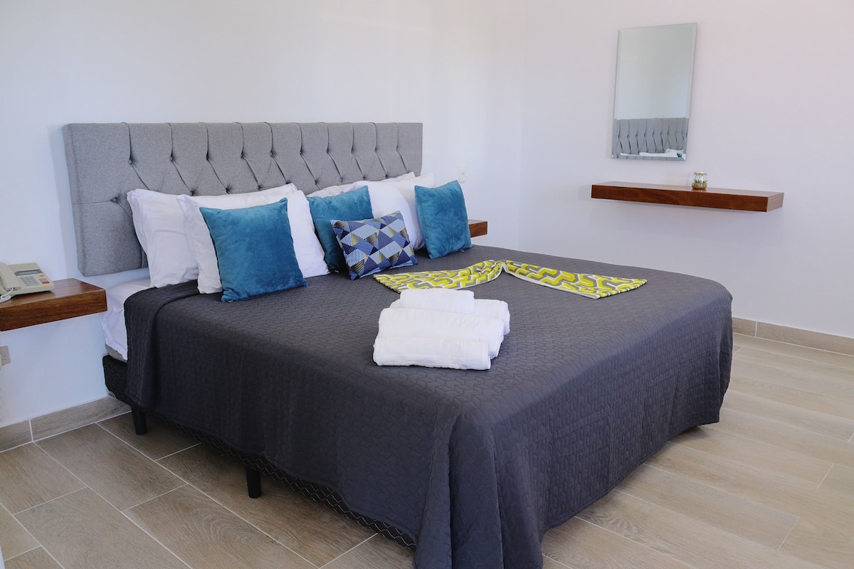 A neatly made bed with a grey headboard, blue and white pillows, and a grey bedspread. There is a wooden nightstand with a telephone and a small plant on a shelf above the bed.