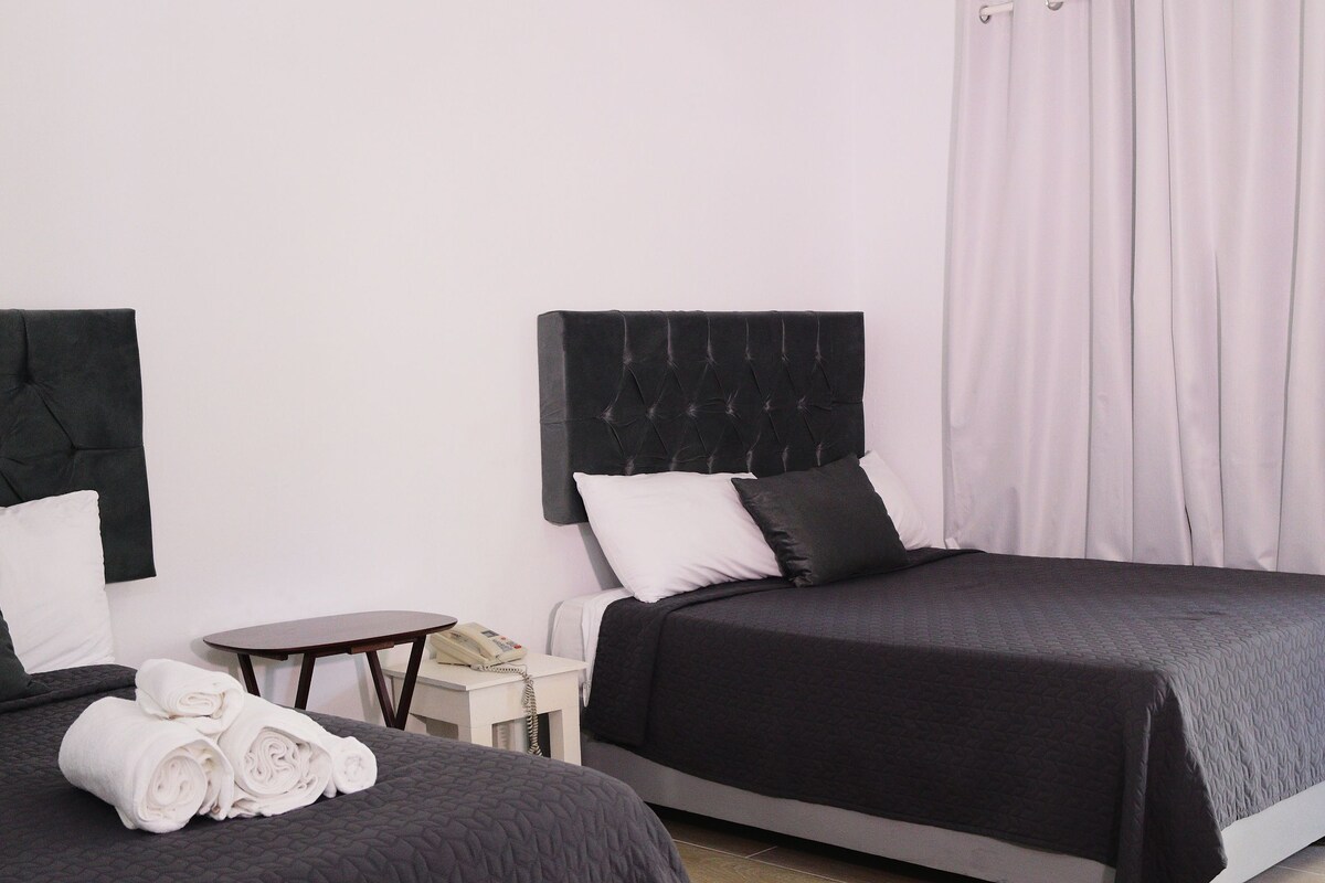 A hotel room with two beds, a small table, a chair, and a bedside table with a lamp.
