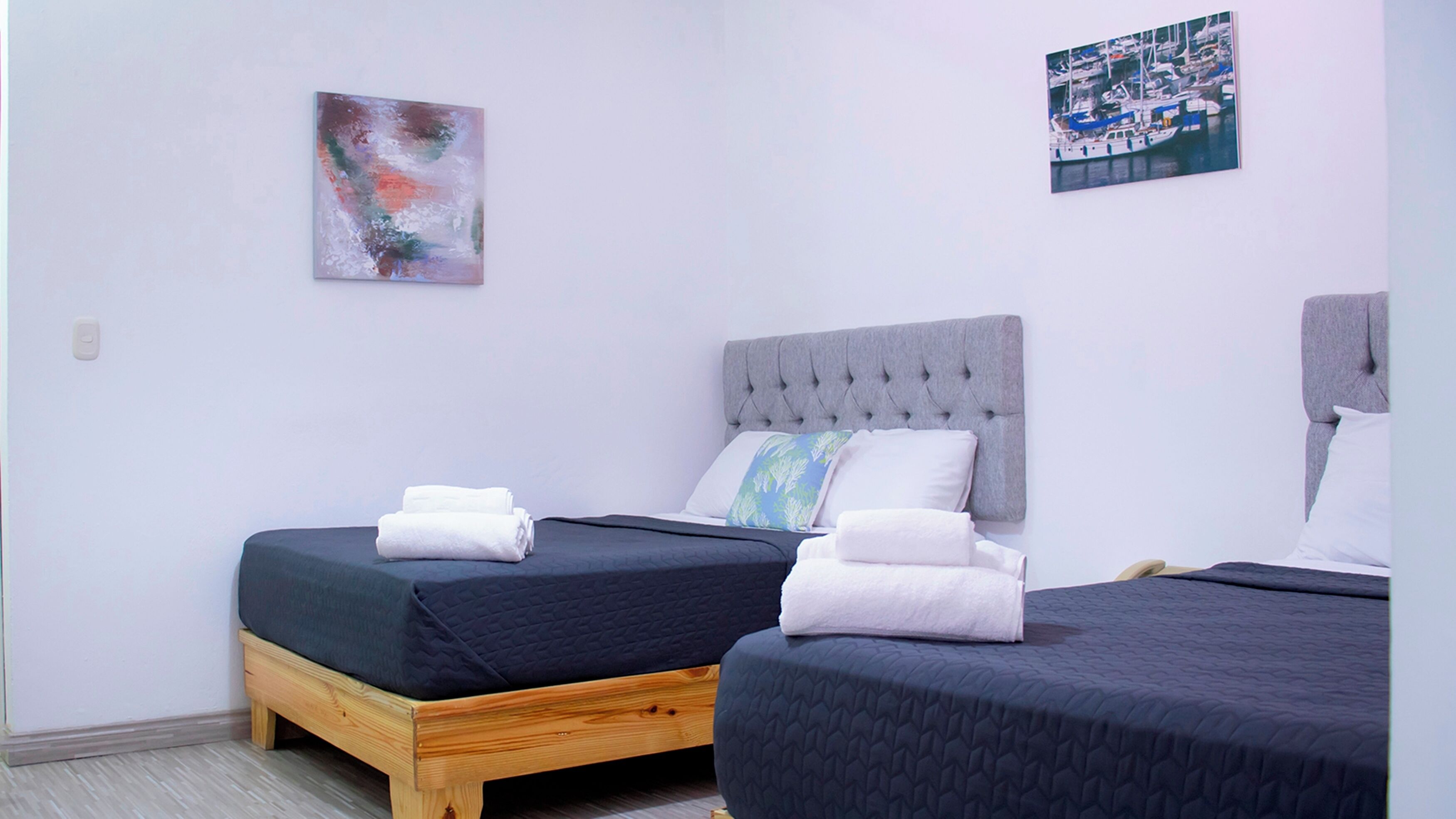 A bedroom with two beds, each with a dark quilted bedspread and white towels. There is a grey tufted headboard on one bed and a wooden headboard on the other.