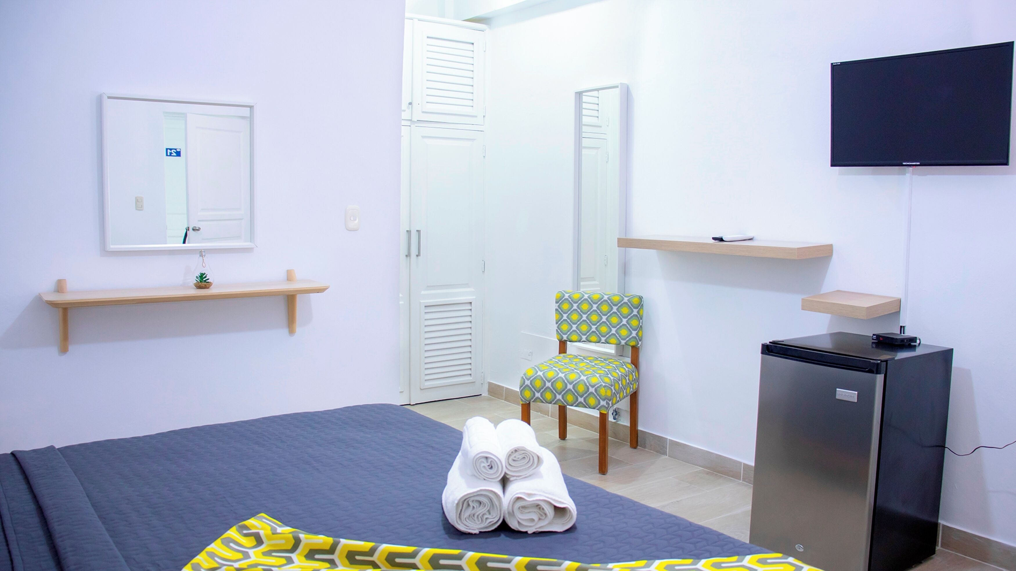 Habitación de hotel con cama, una silla, un escritorio pequeño con computadora, televisor y refrigerador.