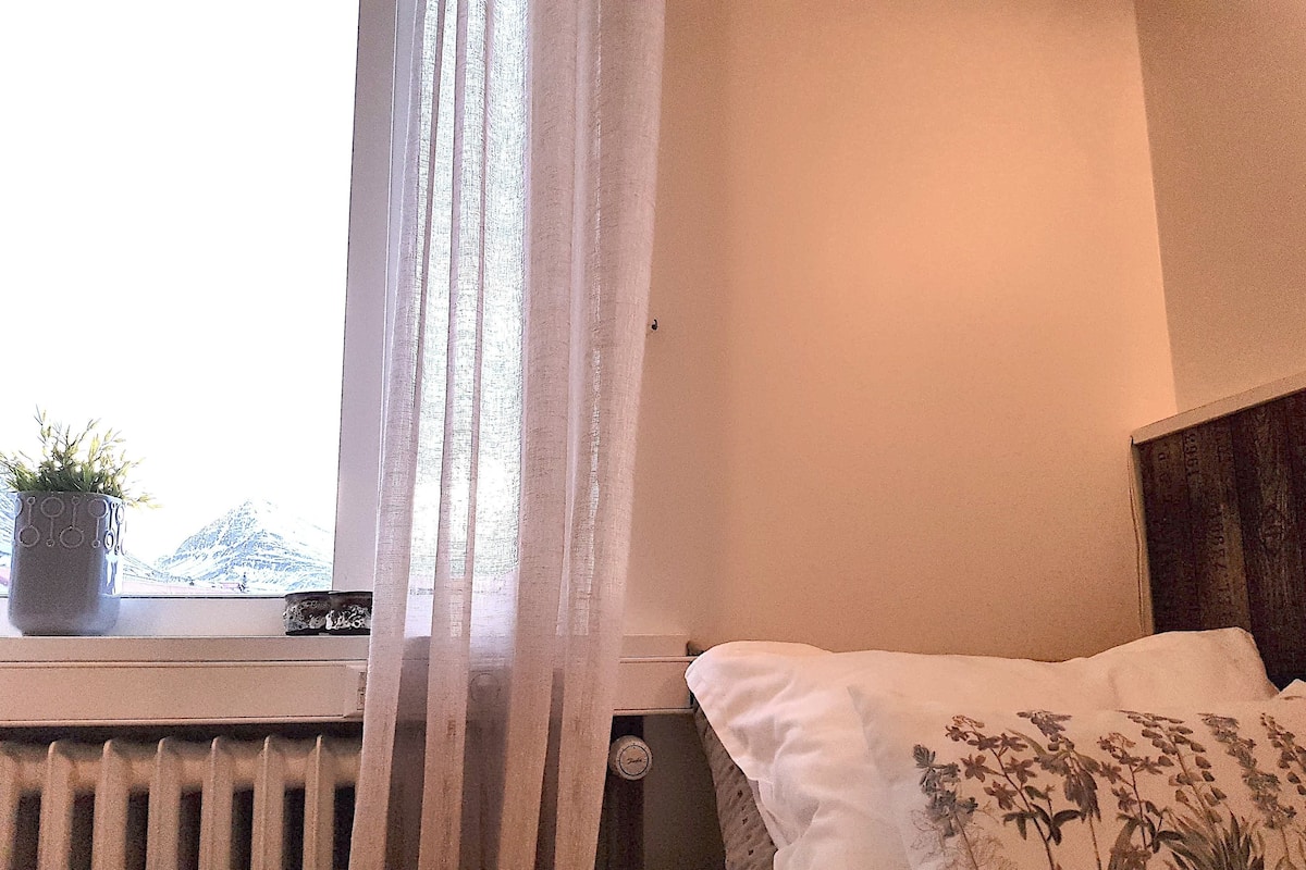 Ein Schlafzimmer mit einem Bett, einem Heizkörper, einem Fenster mit leichtem Vorhang und einem Blumentopf auf der Fensterbank.