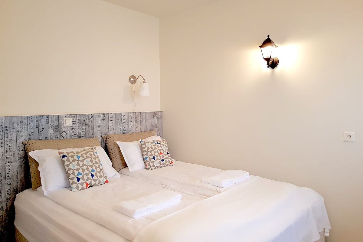 Ein ordentlich bezogenes Bett mit weißen Leinen und gemusterten Kissen, ein Holzkopfschild mit Schriftzug und eine an der Wand befestigte Leuchte.