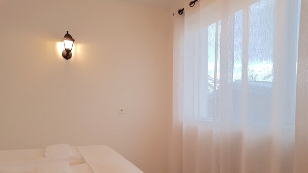 Ein Hotelzimmer mit einem ordentlich bezogenen Bett, einer Wandleuchte und einem Fenster mit durchsichtigen Vorhängen.