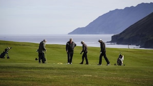 Ein Golfplatz mit Spielern und Golftaschen, mit Blick auf eine Küstenlinie und Berge im Hintergrund.