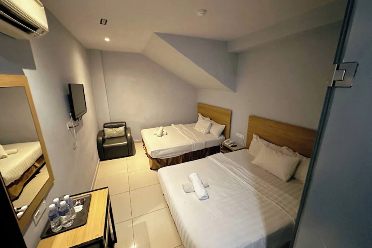 A hotel room with two beds, a small table with water bottles, a TV, and an air conditioning unit.