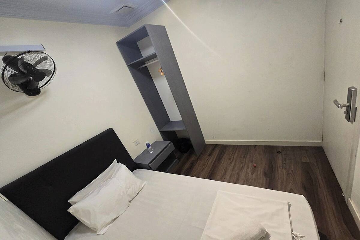 A hotel room with a bed, a small bedside table, a wall-mounted fan, and a built-in shelf unit.