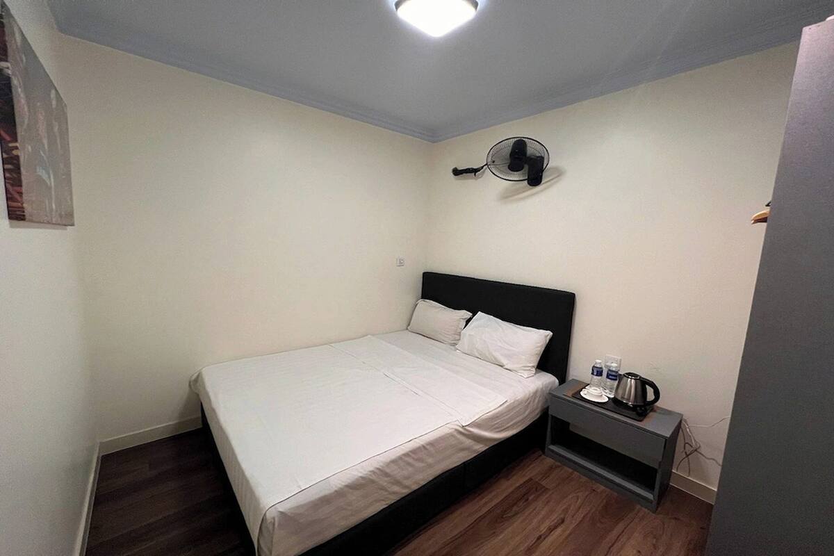 A small, neatly arranged bedroom with a single bed, a bedside table with a kettle, and a wall-mounted fan.