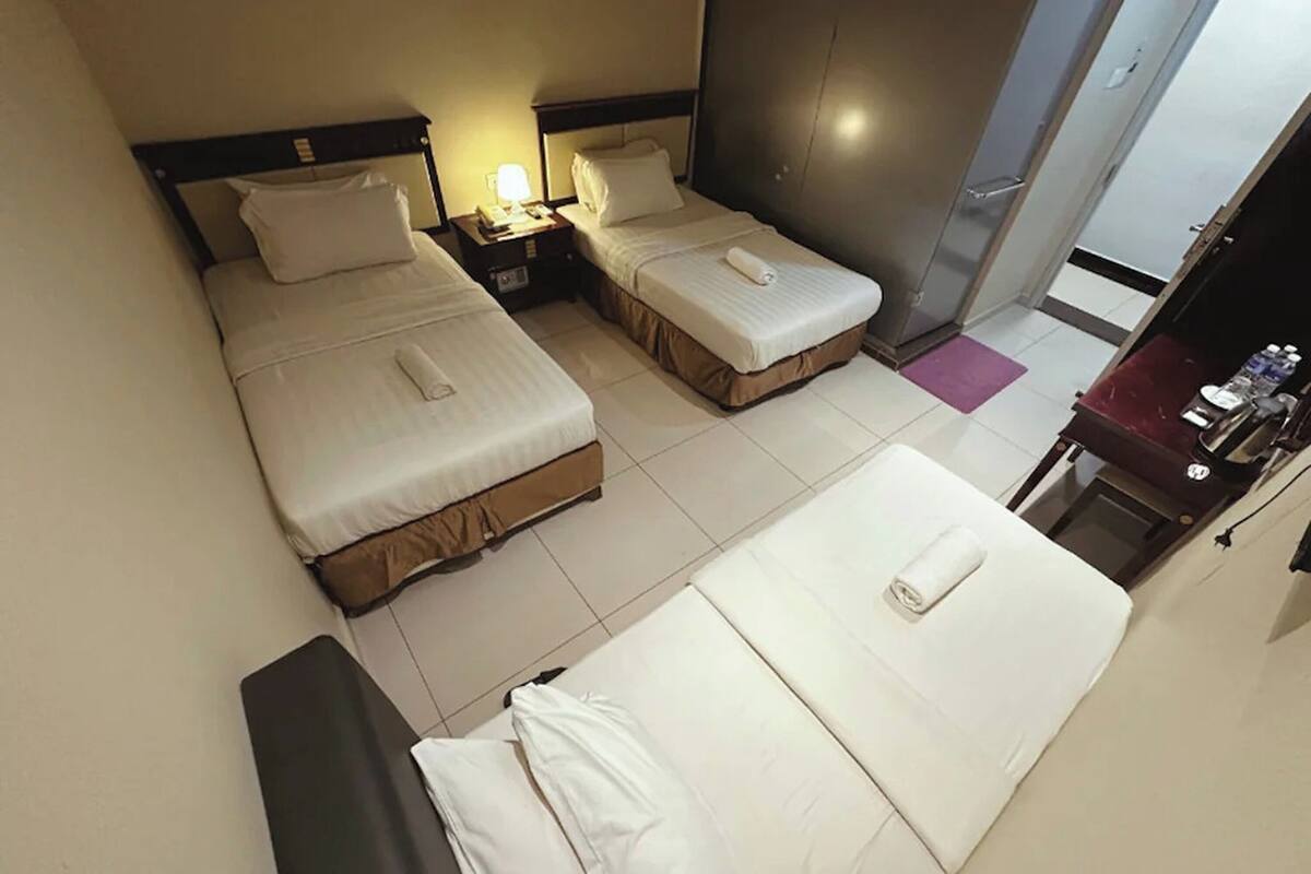 A hotel room with two beds, each with a pillow and a rolled towel. There is a nightstand with a lamp and a phone. The room has a tiled floor and a door leading to another room.