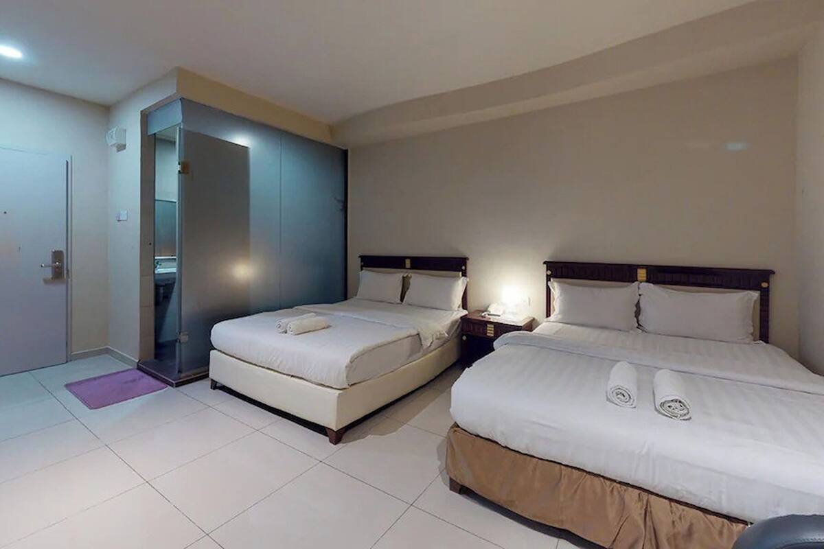 A hotel room with two beds, a bathroom with a glass door, and a small desk with a lamp.