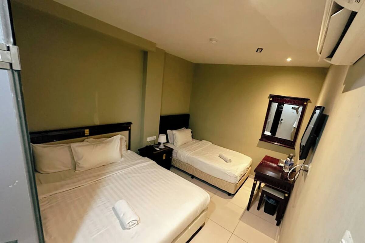 A hotel room with two beds, a bedside table with a lamp, a small desk with a mirror, and a sink.