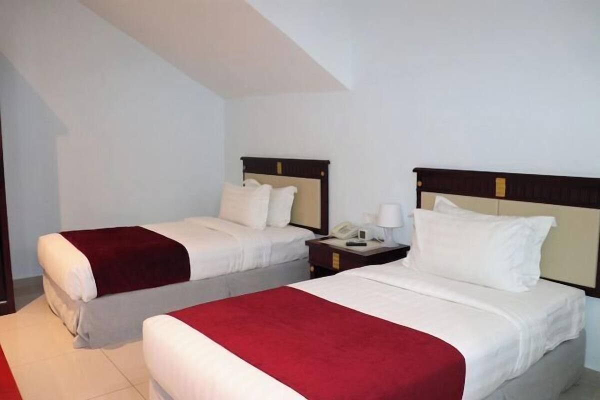 Ein Hotelzimmer mit zwei Betten, jeweils mit weißer Bettwäsche und roten Zierkissen. Ein Nachttisch mit Lampe und Telefon ist vorhanden.
