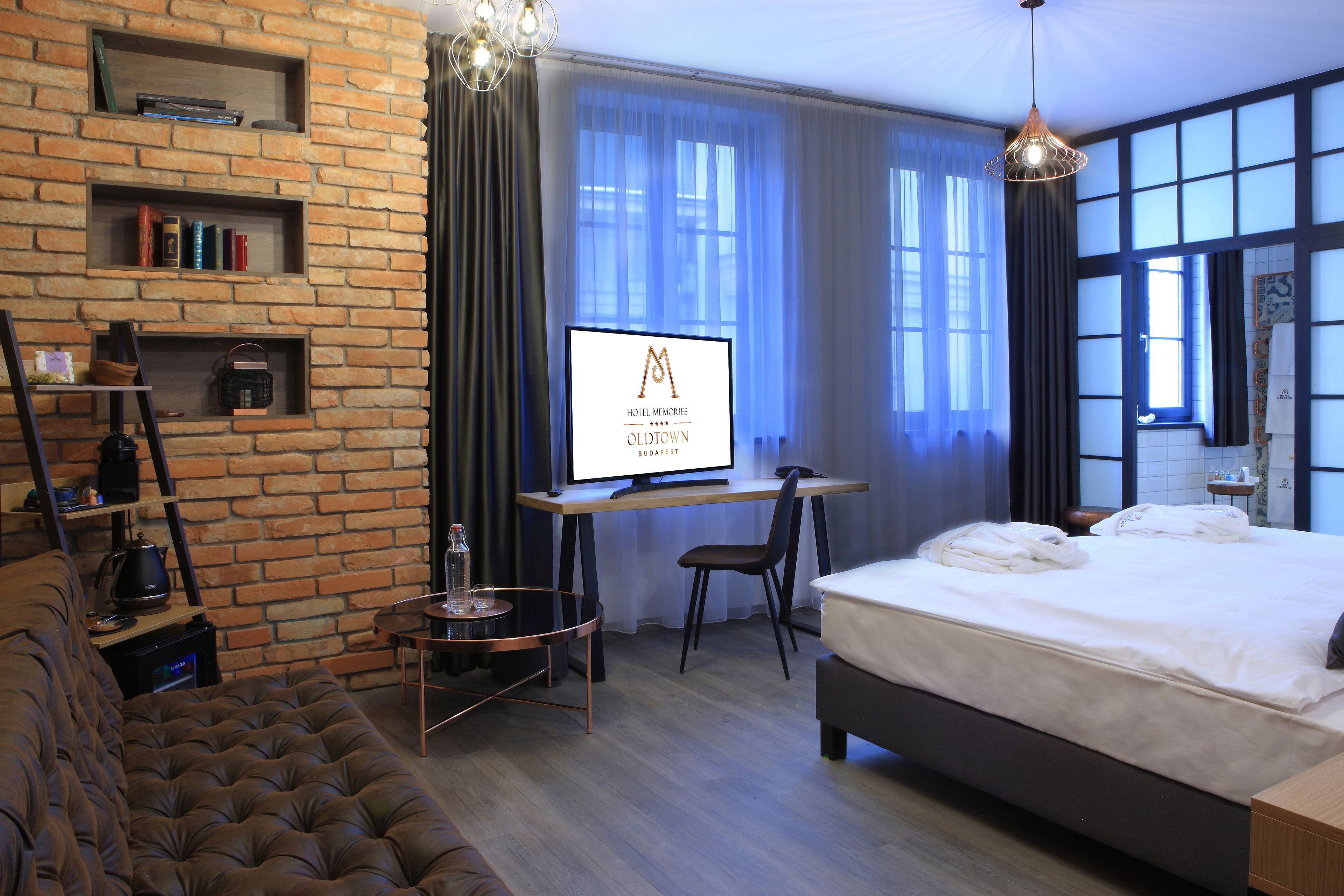 A modern hotel room with a brick wall, a large bed, a desk with a computer, a sofa, and a TV displaying the hotel's name.