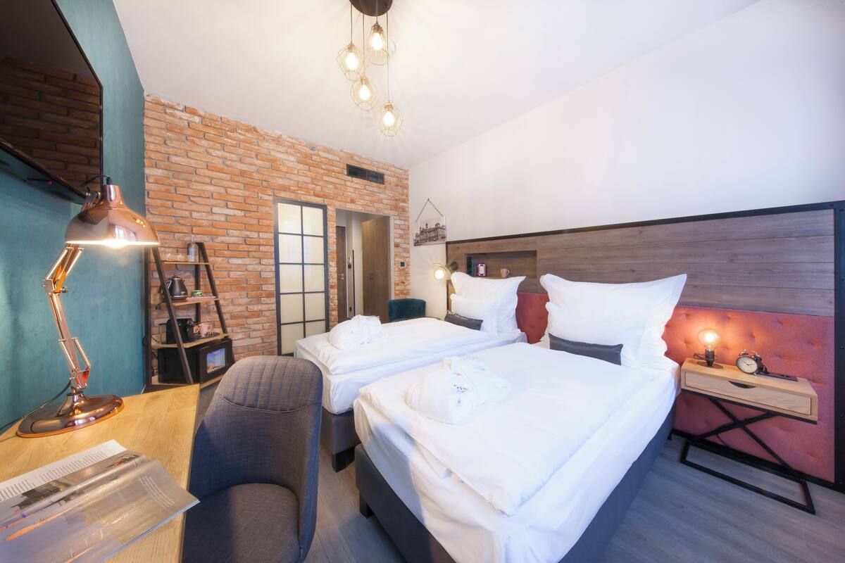 Habitación de hotel con dos camas, un escritorio con lámpara, una silla, pared de ladrillo y cabecero de madera.
