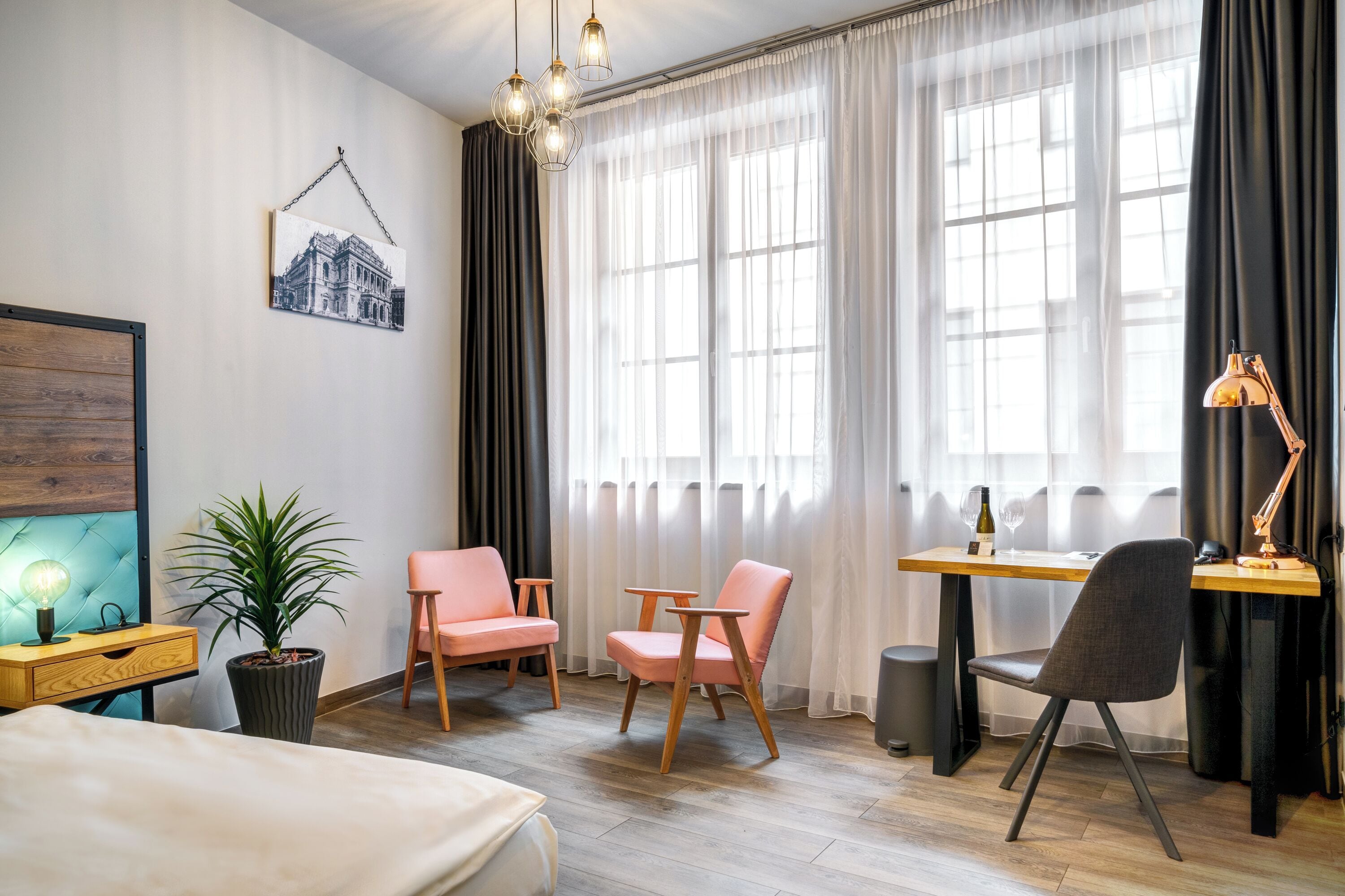 Habitación de hotel moderna con una cama, dos sillas rosas, un escritorio de madera, una computadora portátil, una planta en maceta y un amplio ventanal con cortinas traslúcidas.