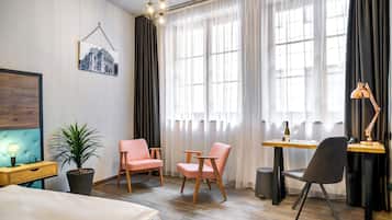Habitación de hotel moderna con una cama, dos sillas rosas, un escritorio de madera, una computadora portátil, una planta en maceta y un amplio ventanal con cortinas traslúcidas.