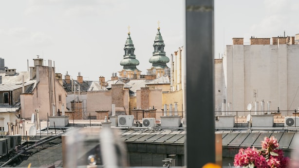 Una terraza en la azotea con una mesa dispuesta para comer, que incluye una copa de vino blanco, un plato de comida y un jarrón de flores. Al fondo, se ven dos torres verdes con cúpulas y un paisaje urbano.