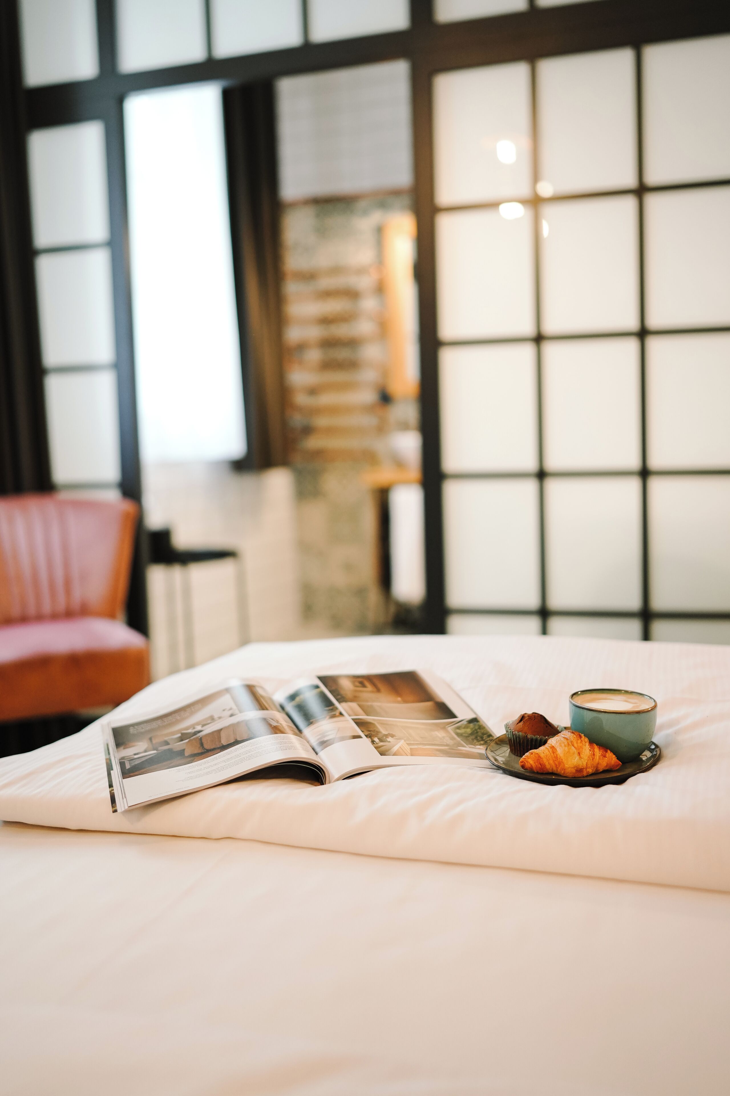 A bed with a magazine, a croissant, and a cup, with a chair and a window in the background.