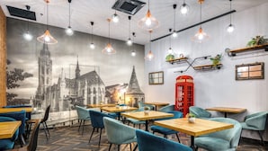 Interior de un restaurante con un mural de una catedral, mesas y sillas de madera, luces colgantes y un teléfono público rojo.